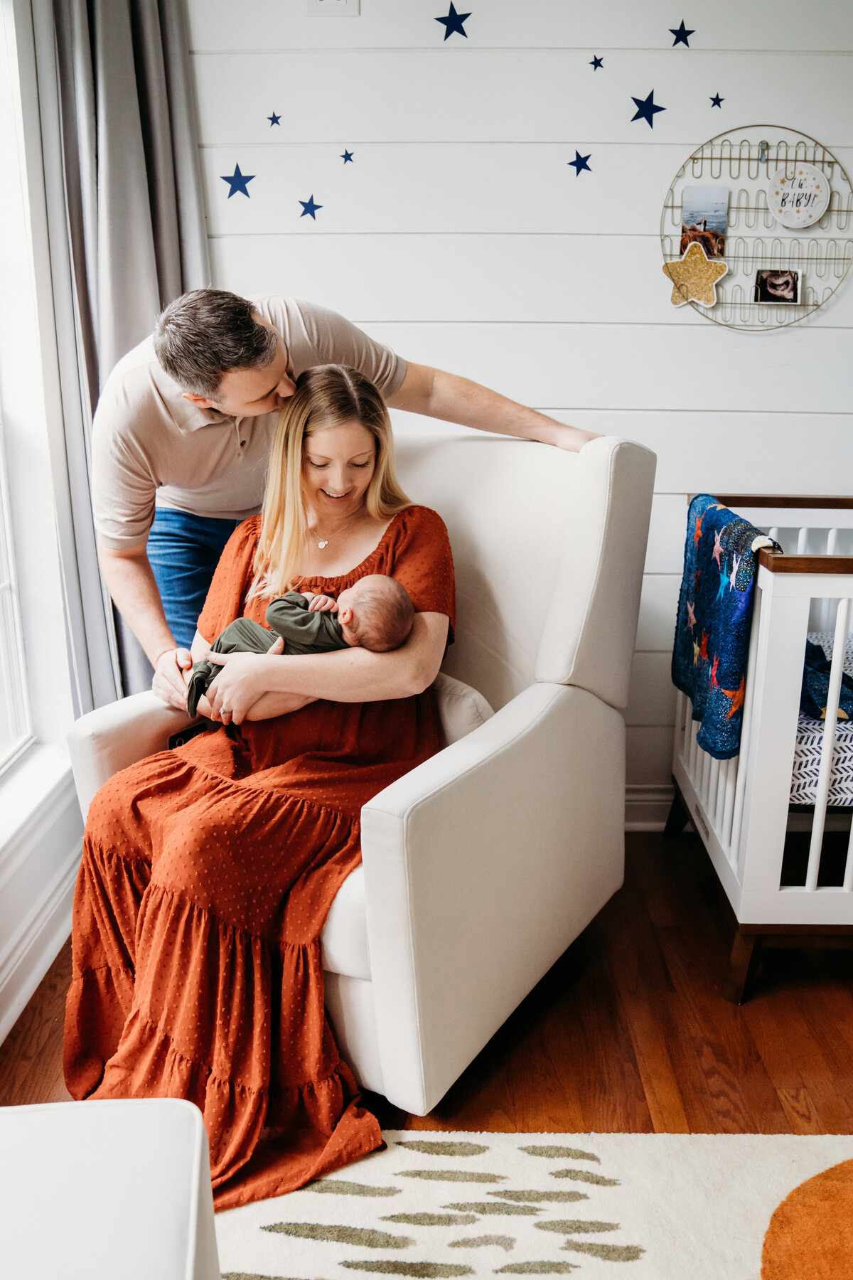 Newborn Photographer, a mother sits in a nursery chair with her baby, dad kisses mom beside them