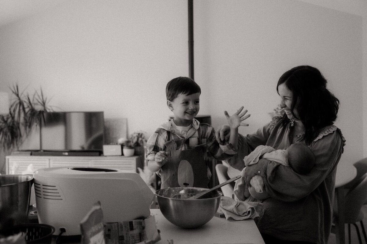 Noir et blanc. Le grand frère rit en montrant ses mains pleines de pâte, sous le regard complice de sa mère qui tient le bébé. Shooting naissance par Kamescop, vidéaste et photographe.