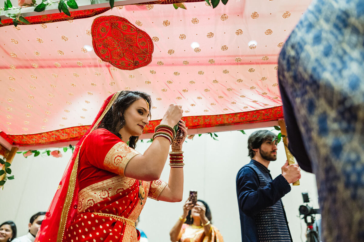 A bride participating in a traditional Indian wedding ceremony under a pink canopy, surrounded by guests.