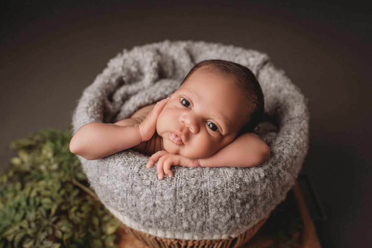 Newborn baby boy looking at camera posed in a basket with hand on cheek