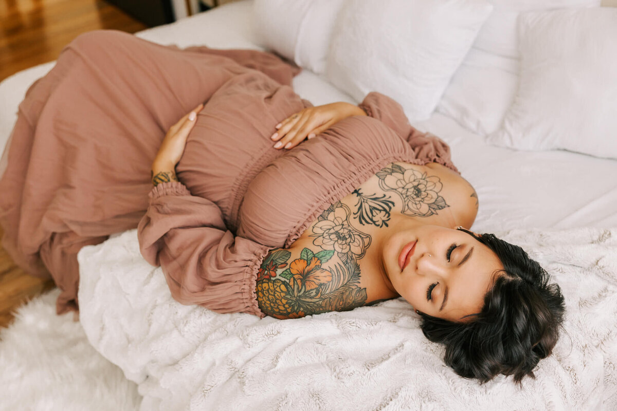 Woman laying on bed with eyes closed. She is holding belly and has her eyes closed. She has black hair and tattoos in studio.