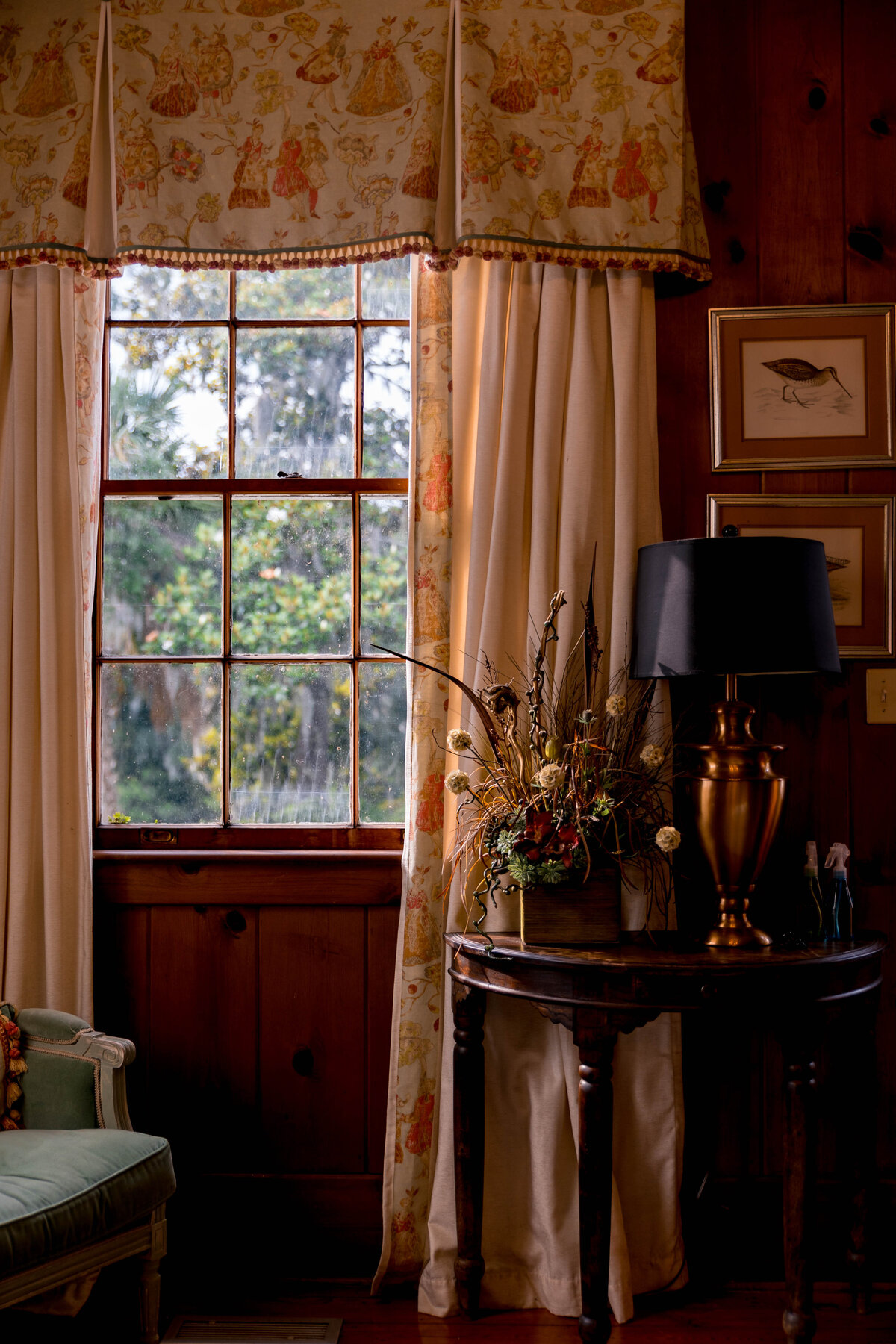 A chair, table, and lamp with a floral centerpiece in front of a window