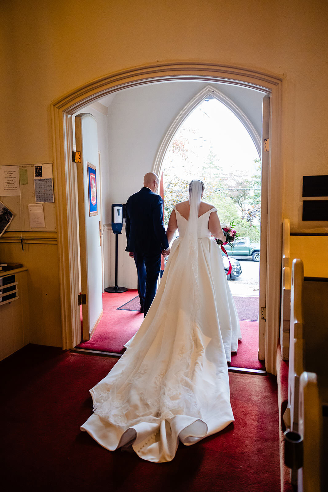 The bride and groom exit a church through a gothic arch door, viewed from behind highlighting the bride's flowing gown