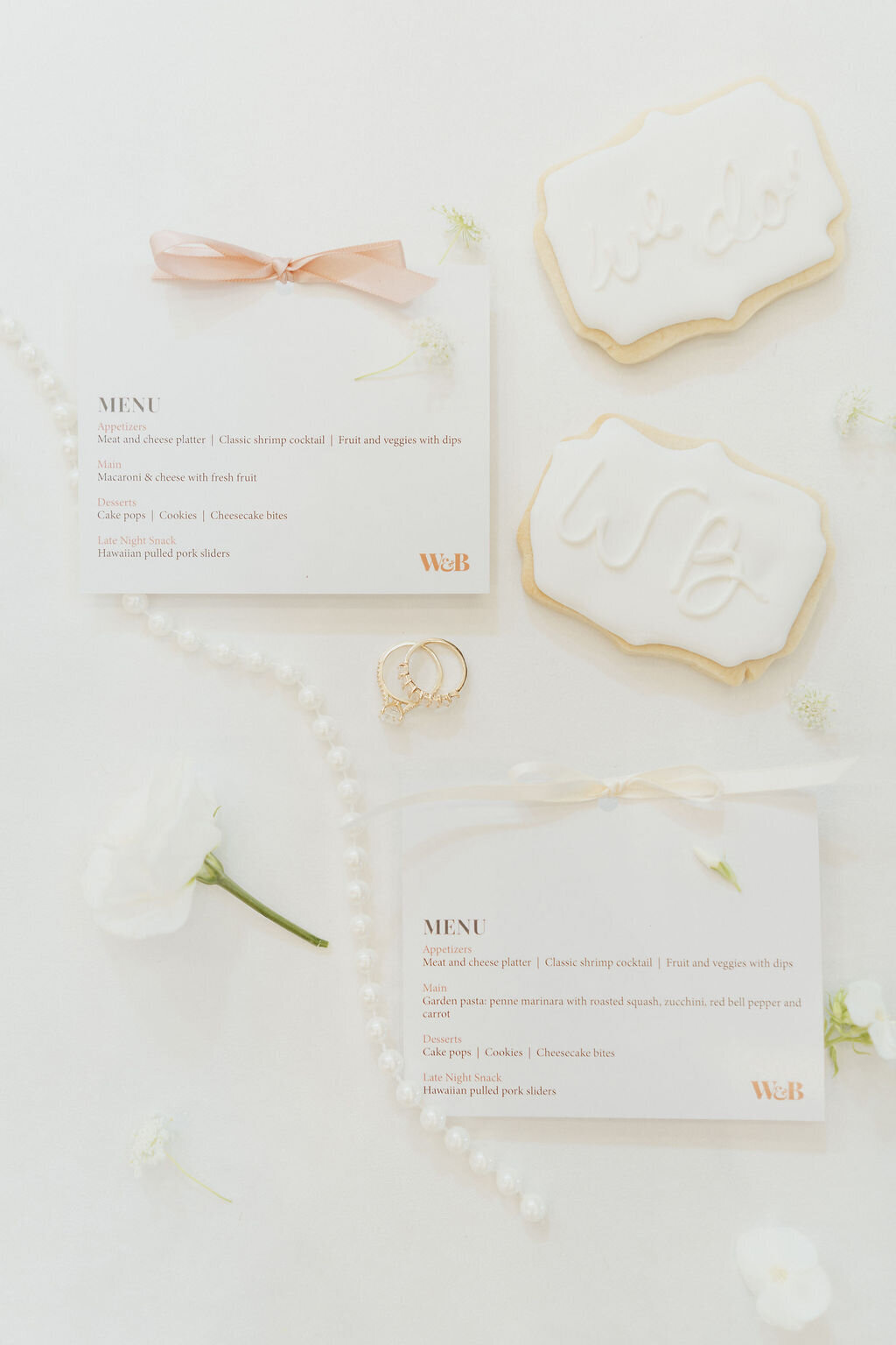 wedding menu cards with accent ribbons placed on a flat lay with monogram cookies, flowers, pearls and rings