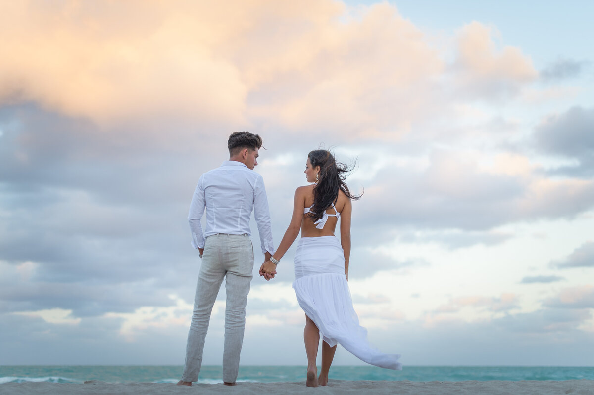 A couple holding hands, gazing out at the ocean under a colorful sunset sky.