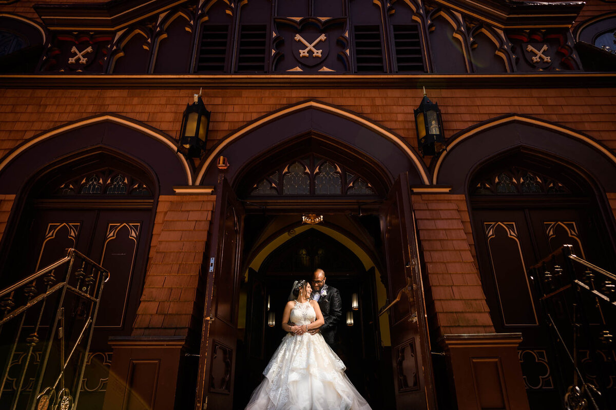 A bride and groom pose together outside of a large building facade.