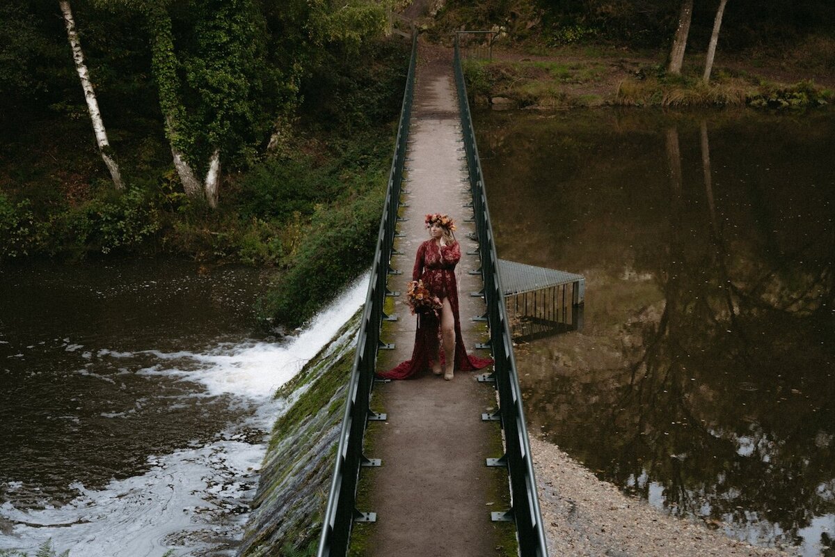 Femme enceinte sur un pont, robe longue en dentelle rouge et couronne florale. Kamescop, séance grossesse artistique en pleine nature.