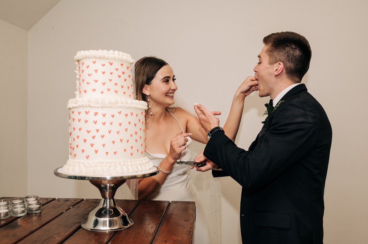 white wedding cake with red hearts in front of bride and groom who are feeding each other a piece at their orton bradley park wedding reception