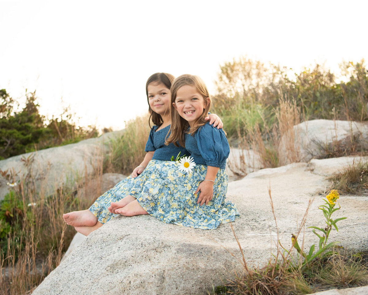 Two sisters in matching dresses sit on a rock at Wingersheek beach, one with a diasy in her hands.