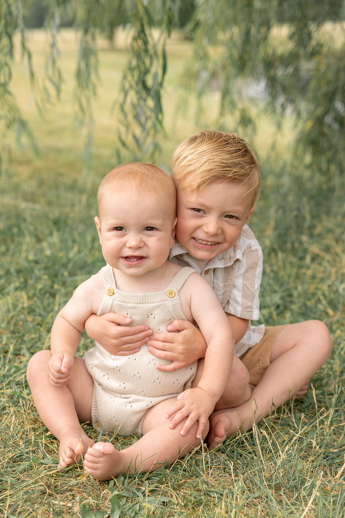 Older brother hugs his baby brother from behind as they smile while sitting in the grass under a willow tree during a session with Indianapolis family photographer, Brittney Lear Photography