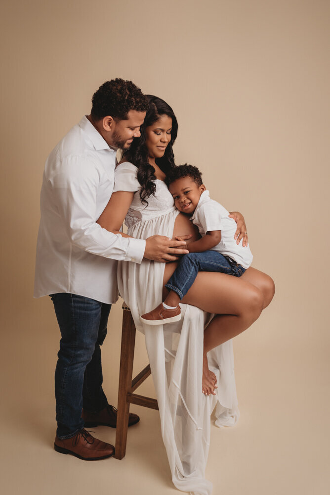 Maternity session with Atlanta, Georgia maternity photographer pregnant woman holding baby bump wearing white sheer maternity  dress on white backdrop posing with husband and 2 year old son  on cream color backdrop