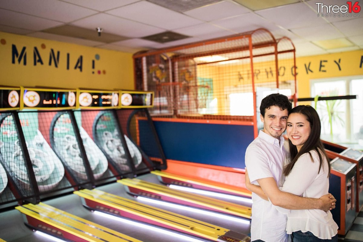 Engaged couple embrace as they pose for photos in front of a skee-ball game