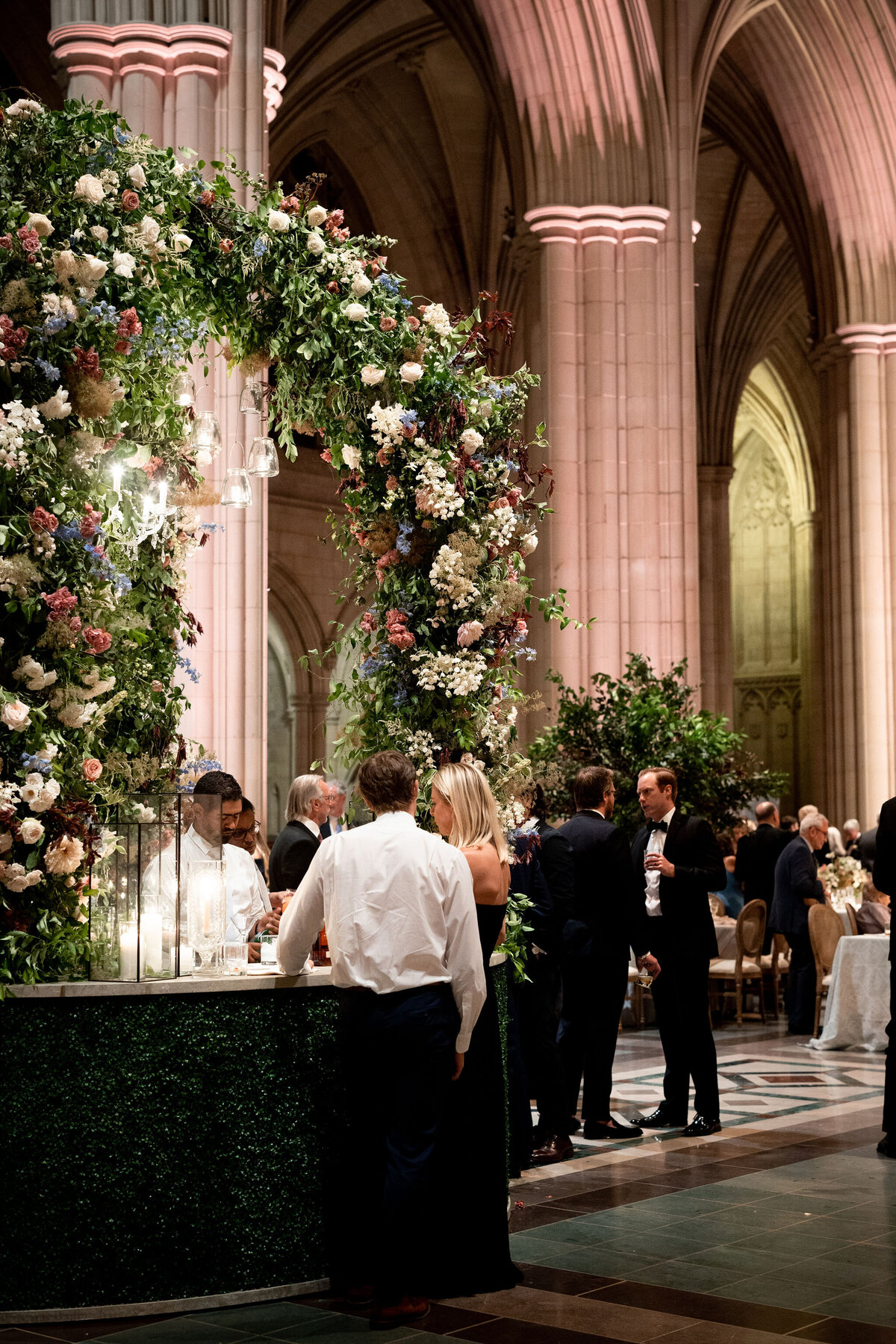 This photo was taken at Washington National Cathedral in Washington D.C. by photographer Twah Dougherty. The grand hall is adorned with floral arches and large columns, creating an elegant atmosphere for a celebration. Guests are seen mingling near a bar, attended by staff, and engaging in conversation at beautifully set tables. The setting exudes sophistication, suggesting a formal event or celebration. If you'd like to add Current Films as videographers to this information, you can mention that they were present at the event capturing the special moments on video.