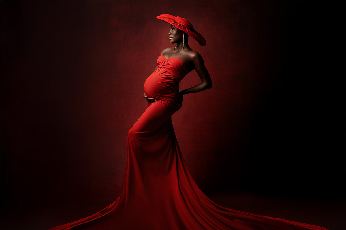 A graceful woman in a stunning red gown and wide-brimmed hat stands confidently against a dark red backdrop, her elegant pose and powerful gaze embodying the artistry of Dallas maternity photographer Lindsay Walden.
