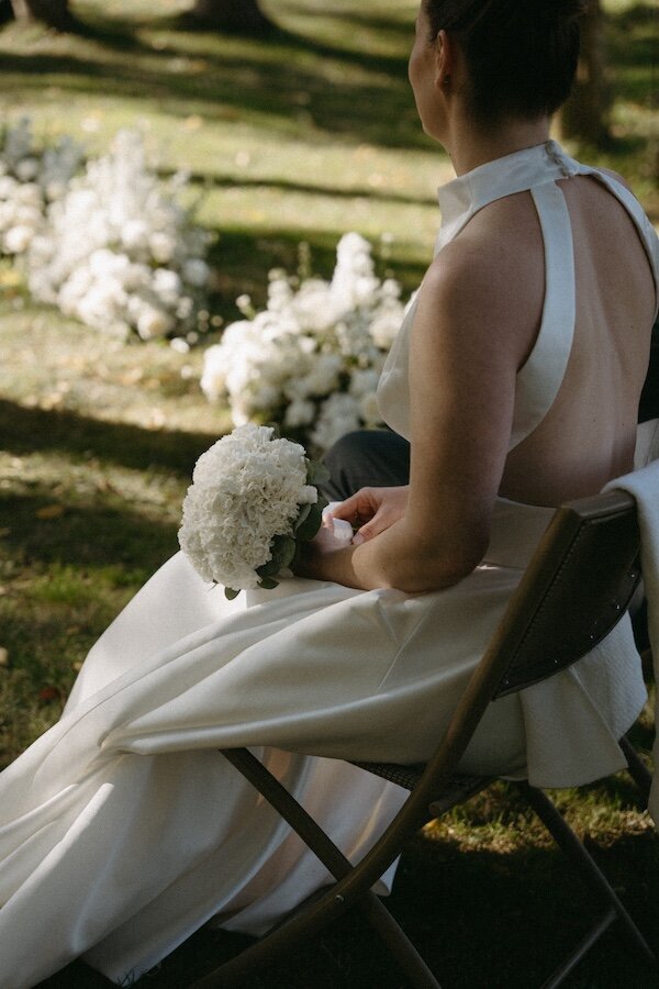 Mariée en robe blanche au dos nu assise pendant la cérémonie, tenant un bouquet d'hortensias blancs. Par Kamescop, photographe et vidéaste mariage.