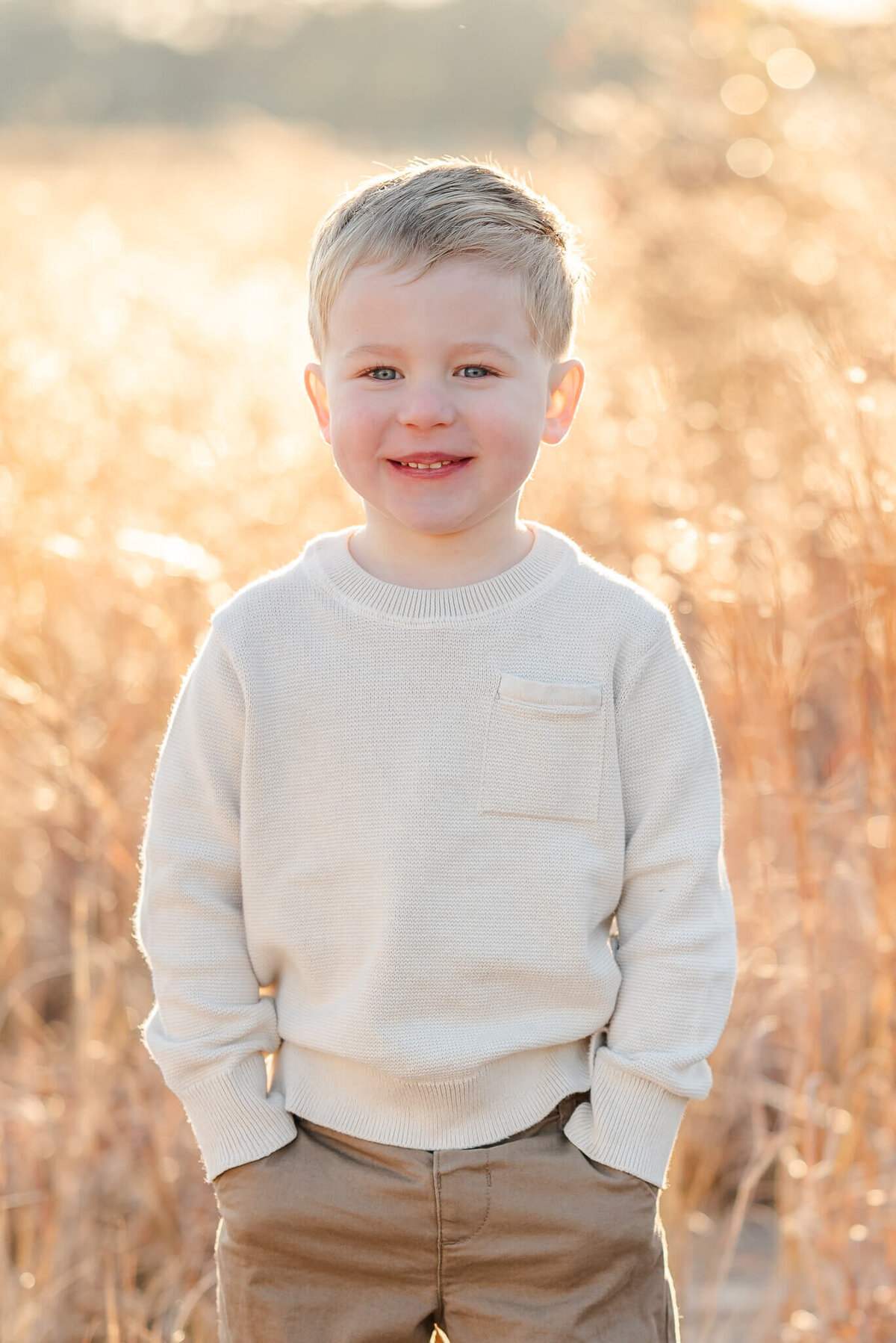 A young boy, wearing khakis and a long sleeve off-white shirt, puts his hands in his pockets and smiles. Photo captured in the Outer Banks by Justine Renee Photography.