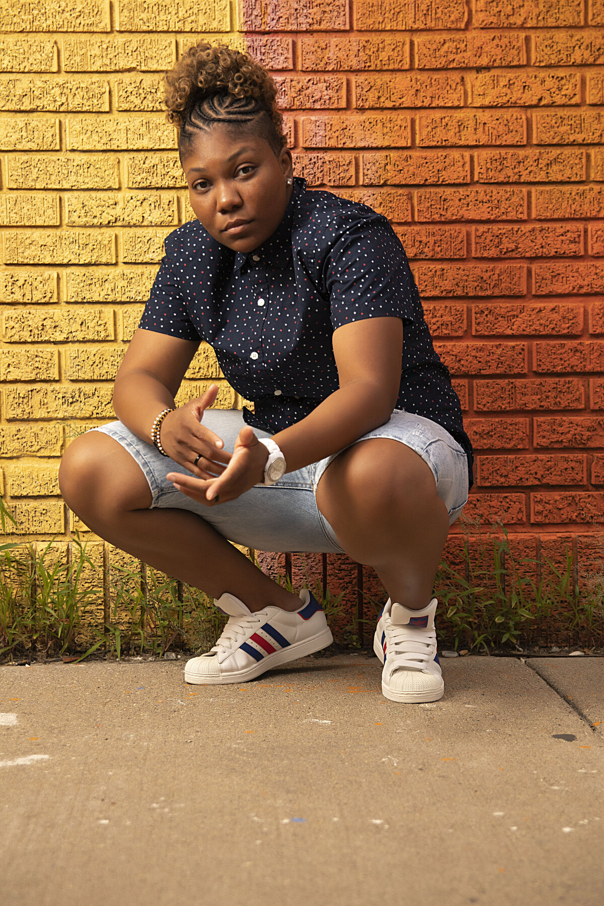 Woman in blue button up top, stonewash blue jeans squatting in front of a colorful brick wall
