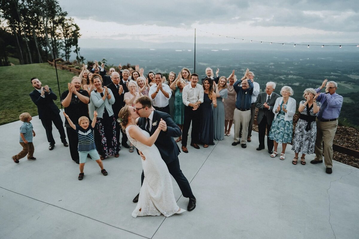 A bride and groom share a dramatic dip and kiss on a patio, surrounded by cheering family and friends