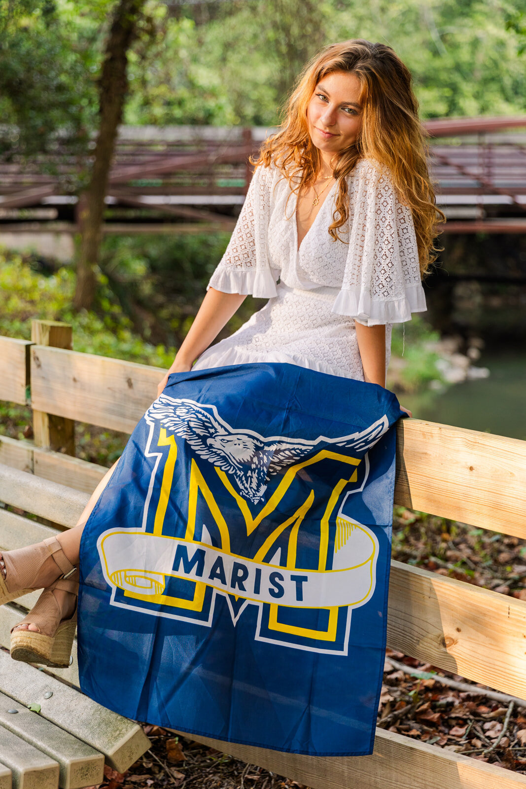 senior girl showing her Marist high school  banner sitting on a wooden fence in front of a bridge