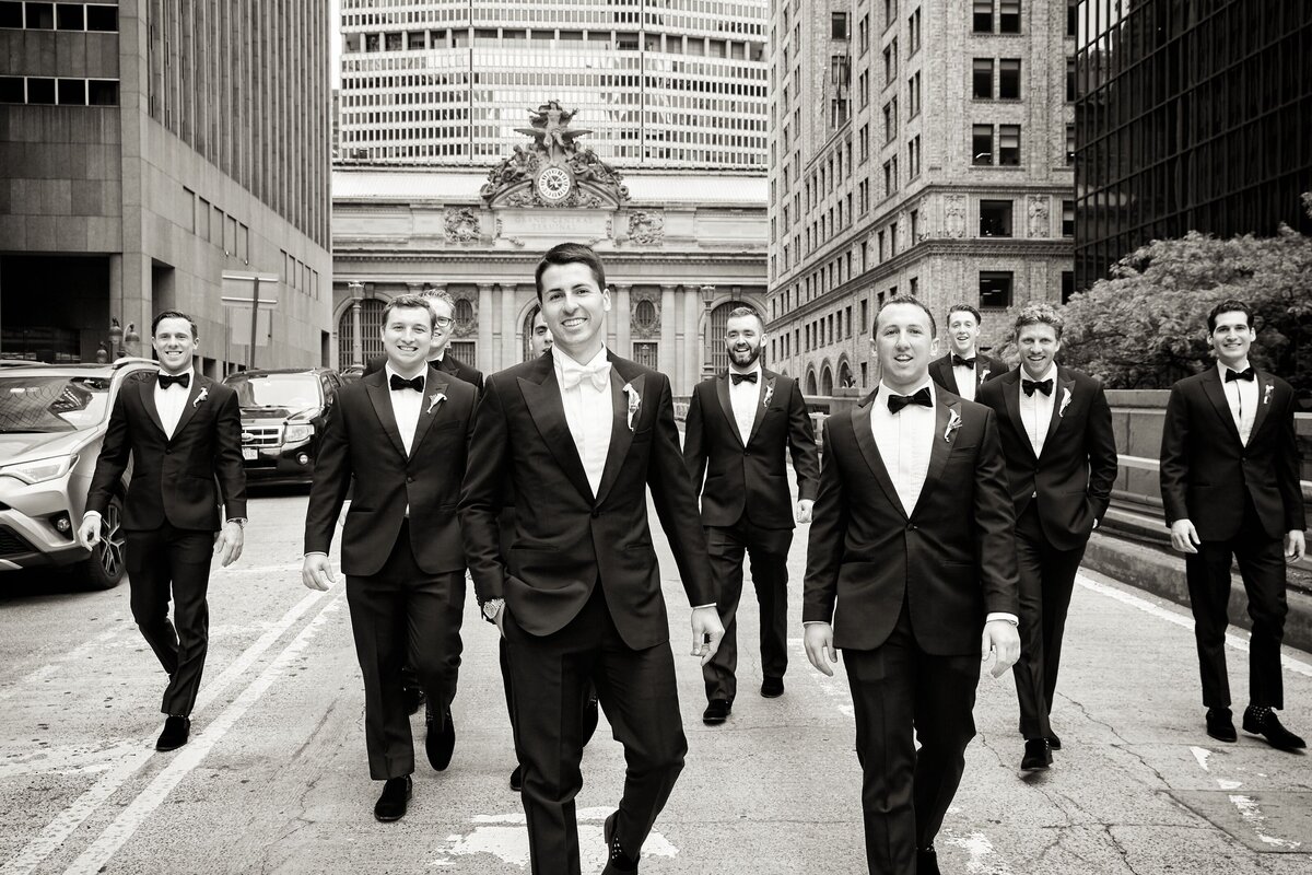 The photo was taken in New York City, specifically at The New York Public Library by photographer Christian Oth. In the image, a group of men in tuxedos, likely groomsmen, can be seen walking confidently down a city street. They appear joyful and are surrounded by tall buildings. The backdrop features a grand architectural structure with a large clock, adding a touch of elegance to the scene. The photo is captured in black-and-white, adding a timeless and classic feel to the overall composition. East West Films can also be credited as the videographers.