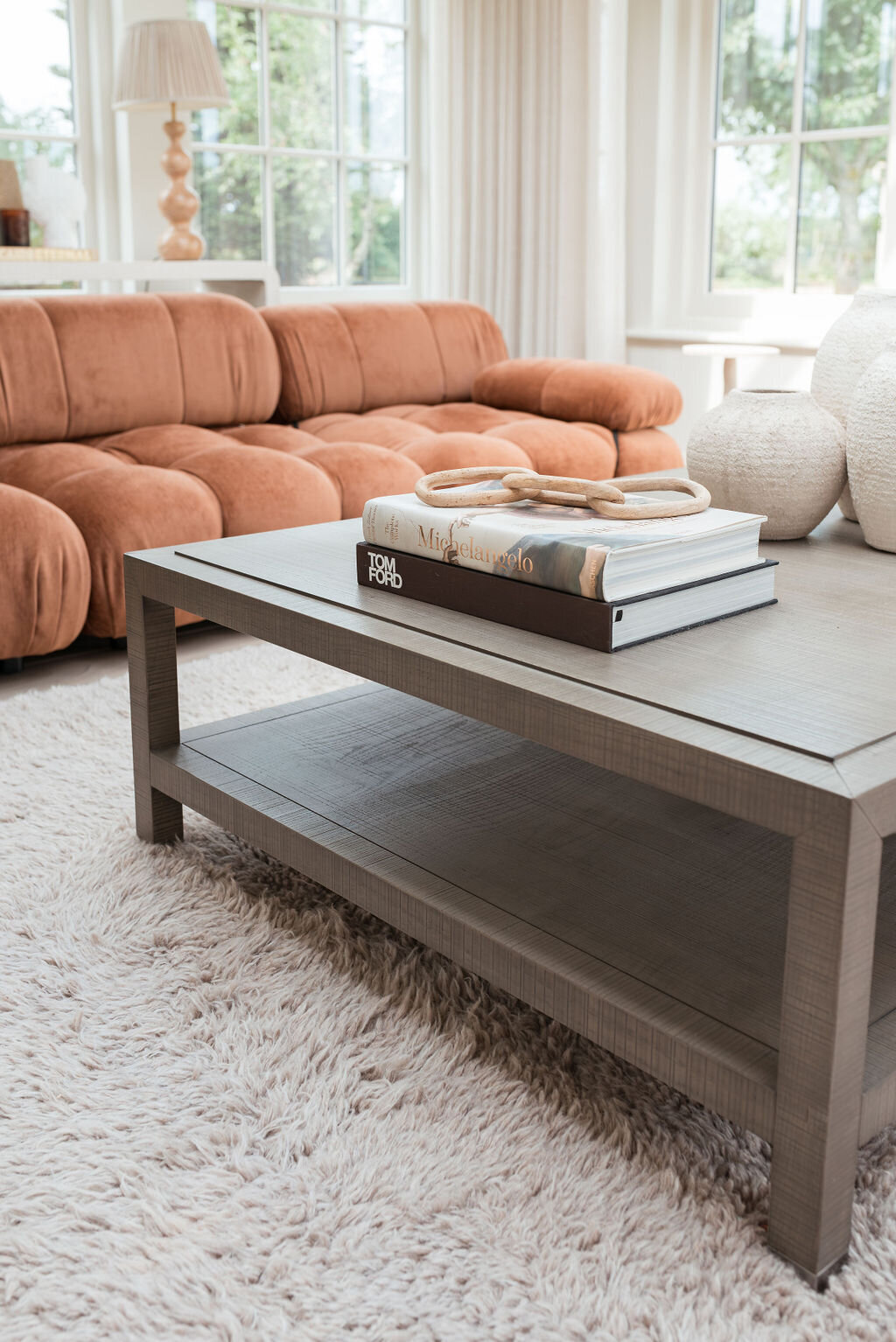 A brown coffee table atop a neutral textured rug. On the table are books and chic decor. Behind the table is a large, plush orange couch.