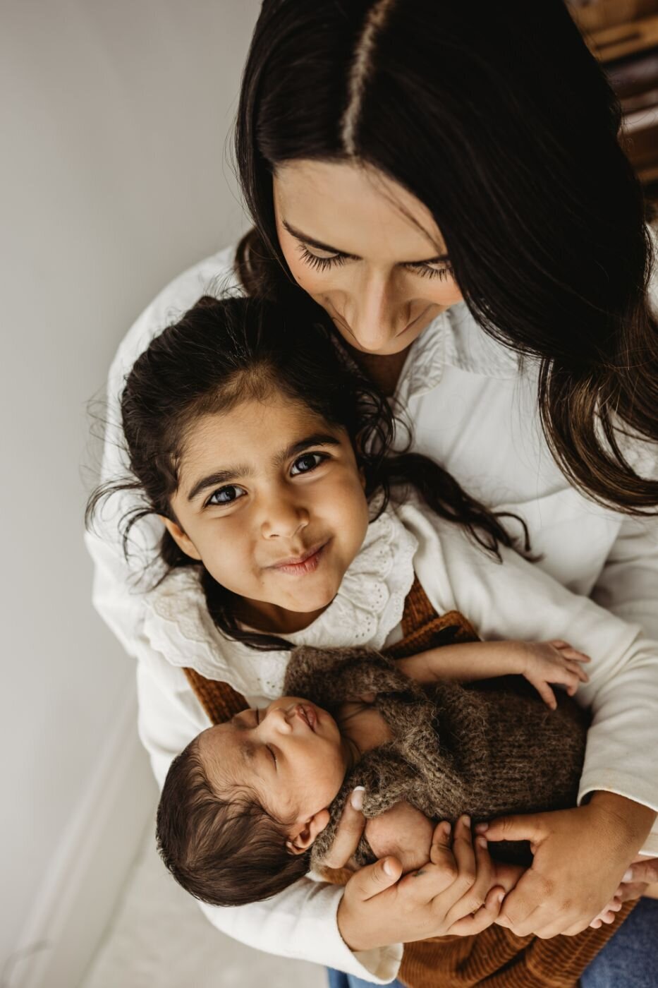 A mother holds her newborn and older daughter. The older daughter looks directly at the camera and smiles.