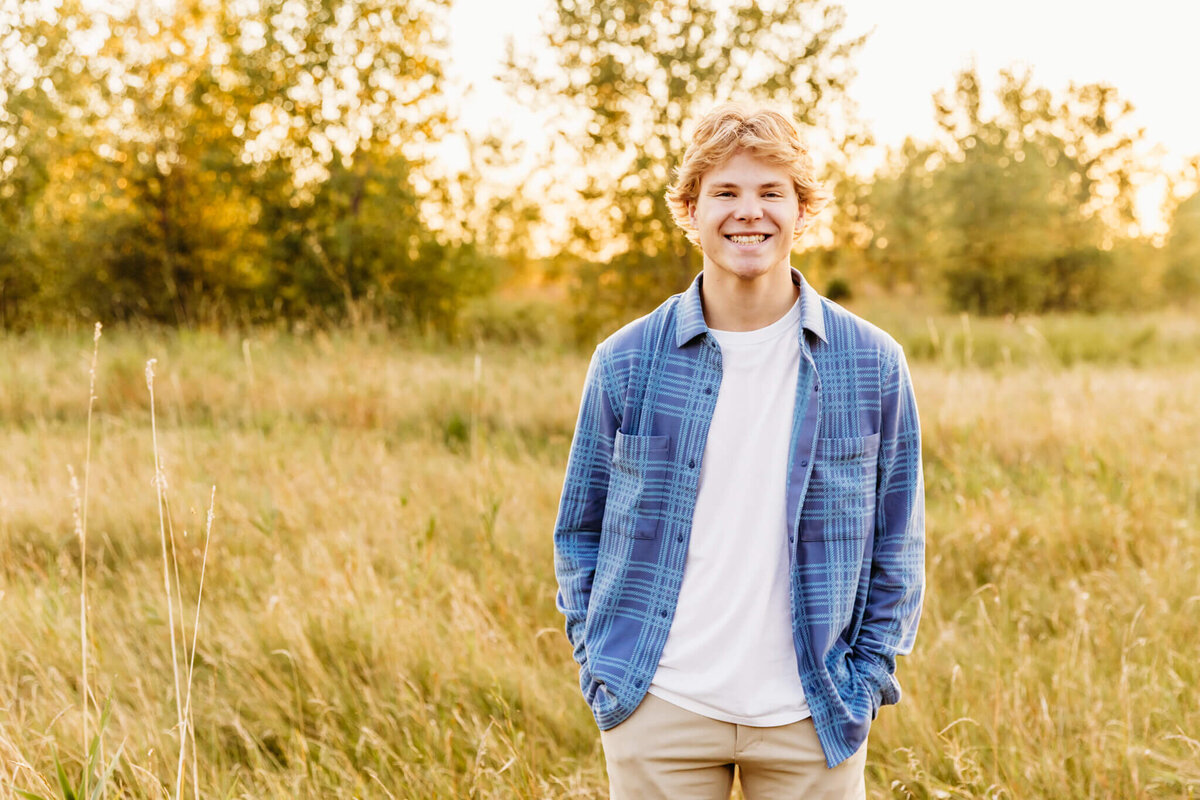 male high schooler with a white shirt and blue flannel smiling as he stands in a field at sunset with his hands in his pockets captured by Neenah senior photographer Ashley Kalbus.