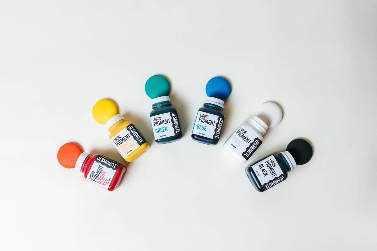 Six different colored paints are laid in an arch on a white table with puddles of paint above each.
