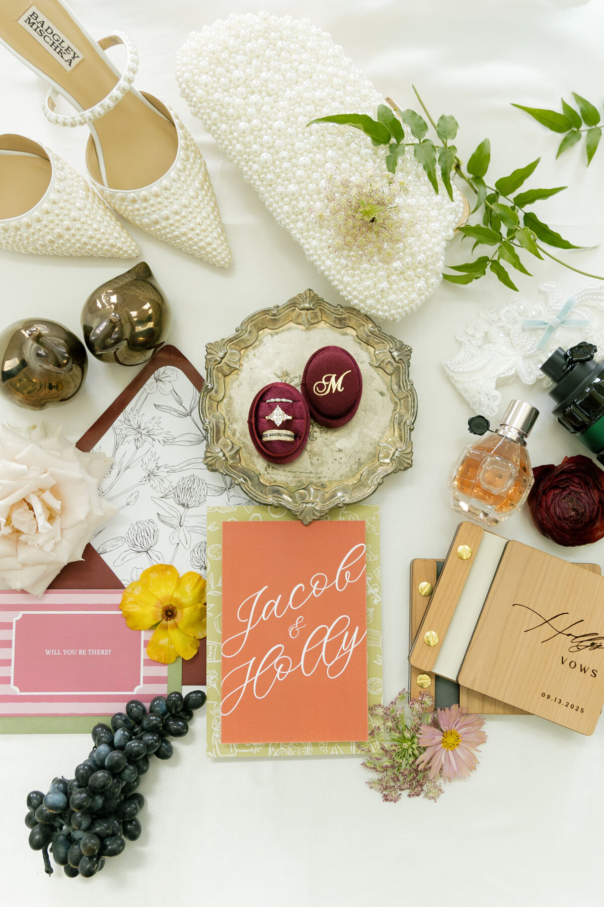 A luxury wedding flatlay featuring pearl-studded bridal heels, a pearl clutch, invitation suite, vow books, yellow ranunculus, perfume bottle, grapes, lace accessories, and a velvet ring box with diamond wedding bands. Styled on a soft ivory backdrop for a romantic, editorial bridal aesthetic.