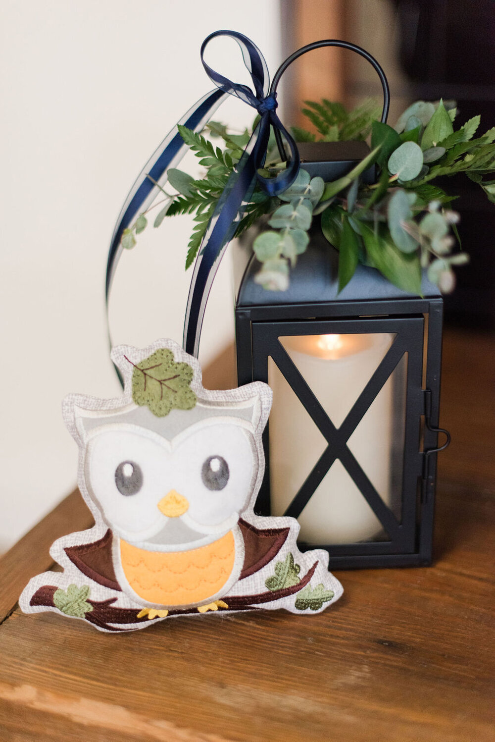 A decorative lantern with a candle, a charming owl figurine, and greenery arranged on a wooden cart for a baby boy shower.