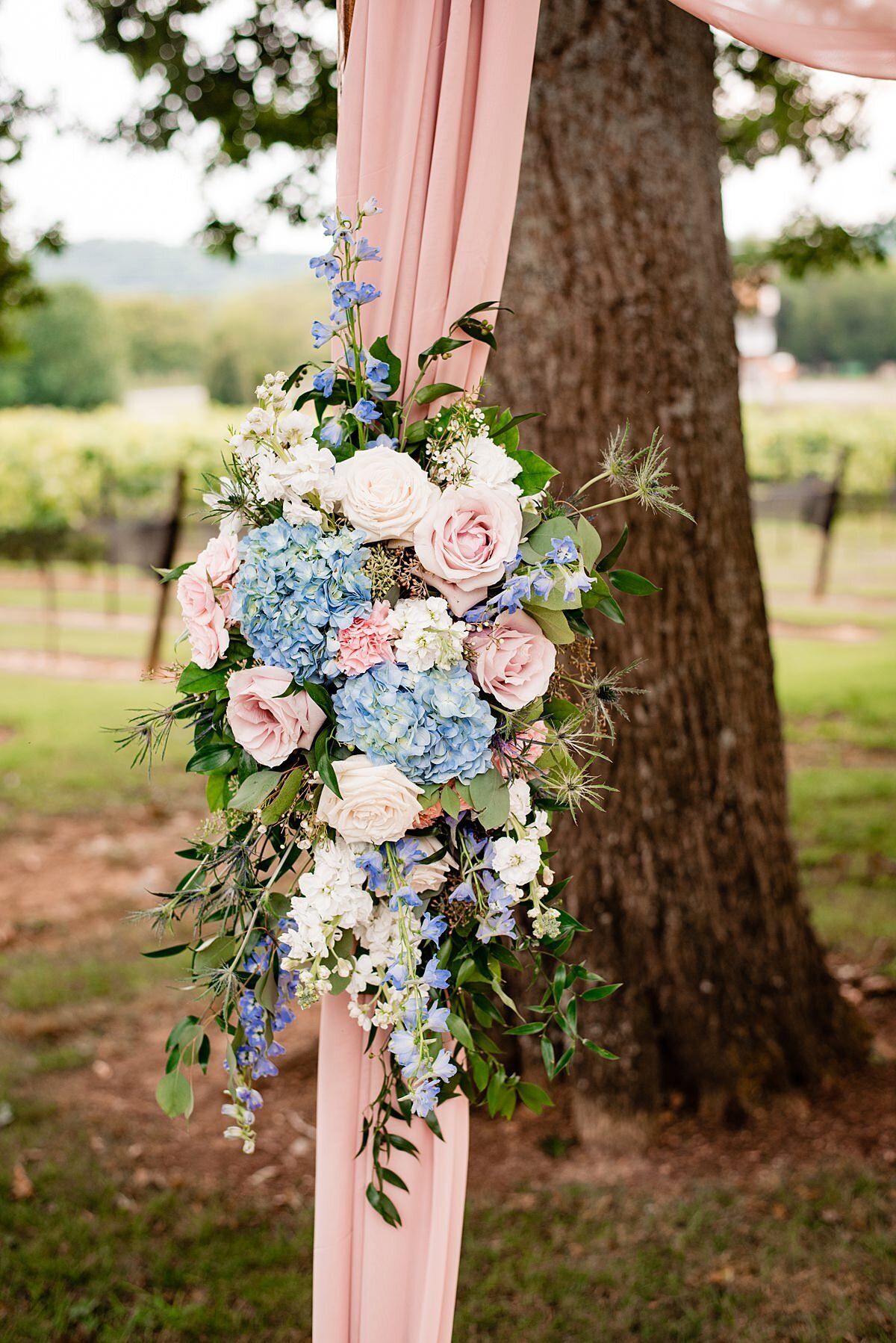 Wedding arbor decorated with sheer blush pink fabric and a large spray of blue hydrangea, blush roses, white roses, white hydrangea, greenery and eucalyptus.