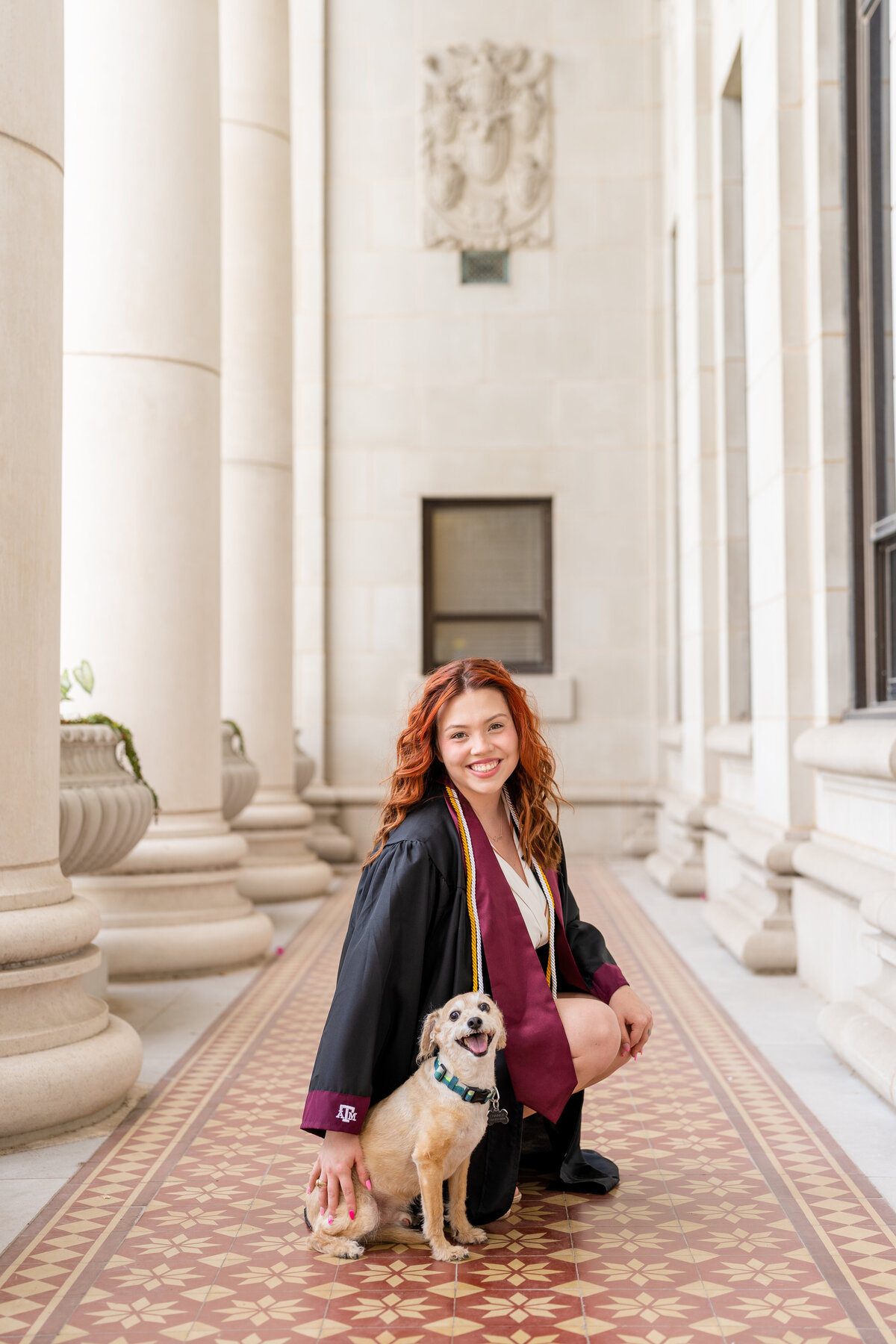 Texas A&M senior girl squatting down next to her dog and both smiling at camera while wearing gown, cords and stole in middle of columns of Administration Building