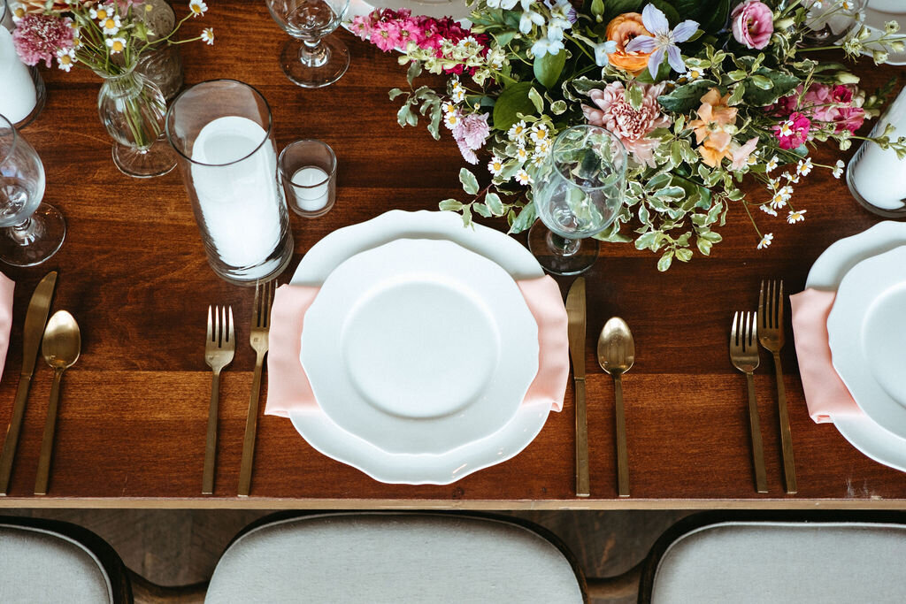 A tabletop set with white china and brass flatware and candles and pink peach and white wildflowers at Willowbrook wedding venue