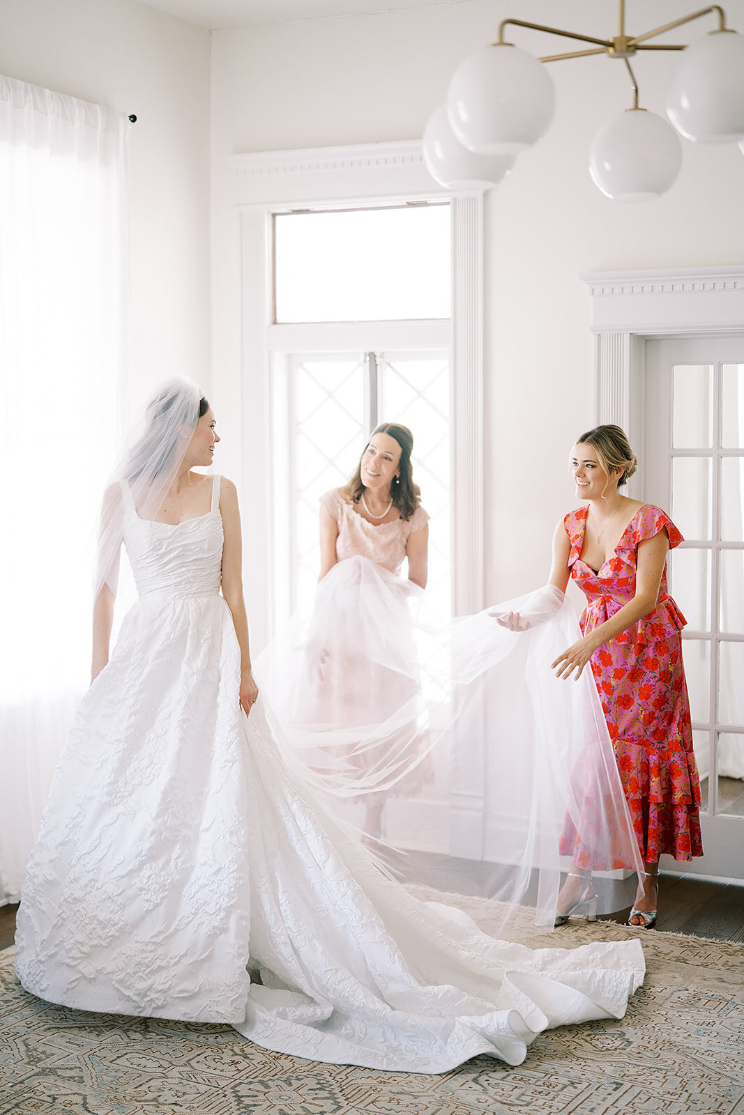 The bride's mother and sister help her with her veil in the brightly lit bridal suite
