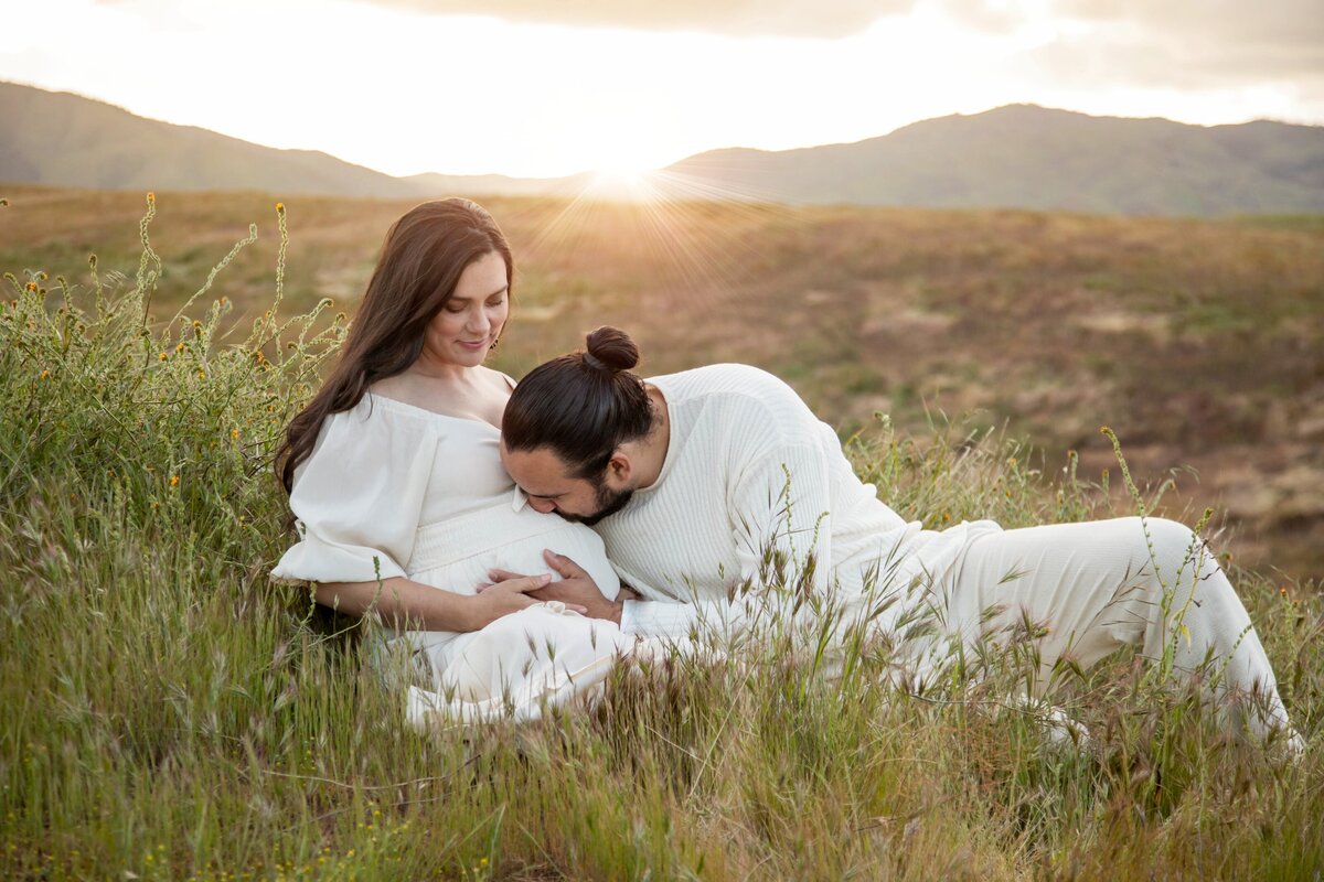 Couple sitting in field, expecting father kissing belly | California Maternity Session