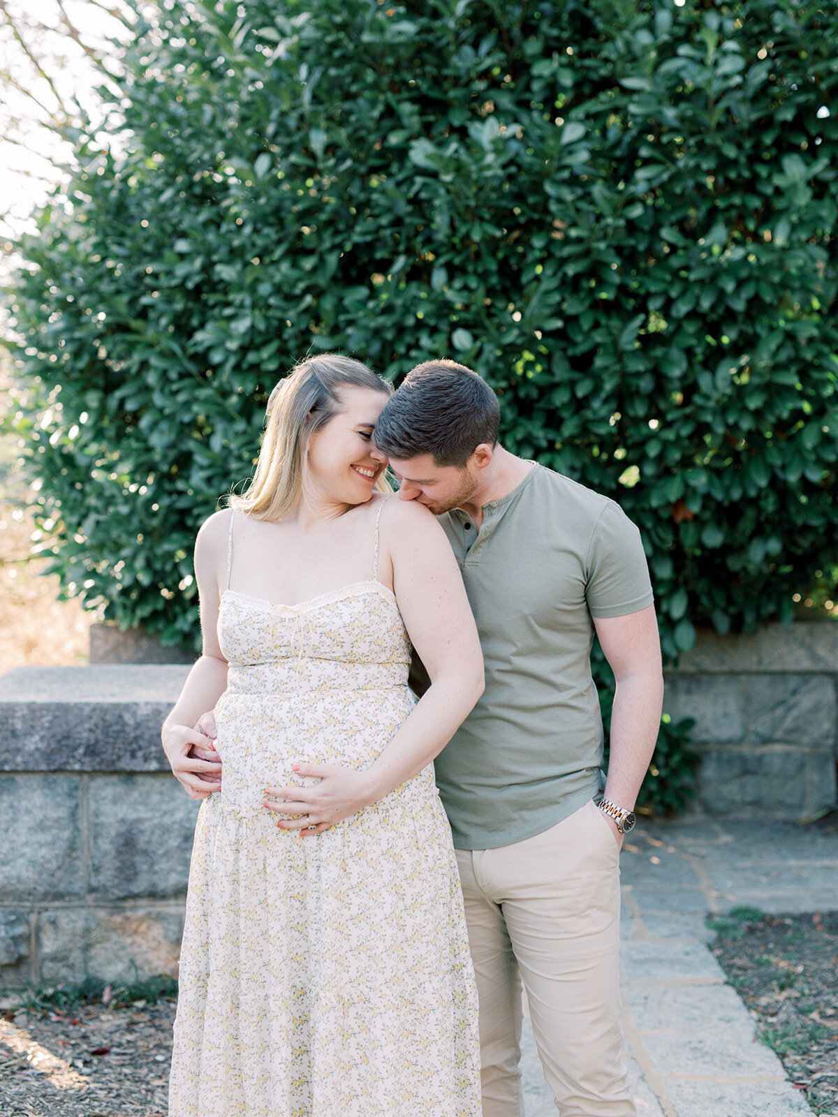 A man kissing a woman's shoulder with the woman smiling at him and holding her pregnancy bump at a Richmond maternity photography session by Katie Stansfield Photography.