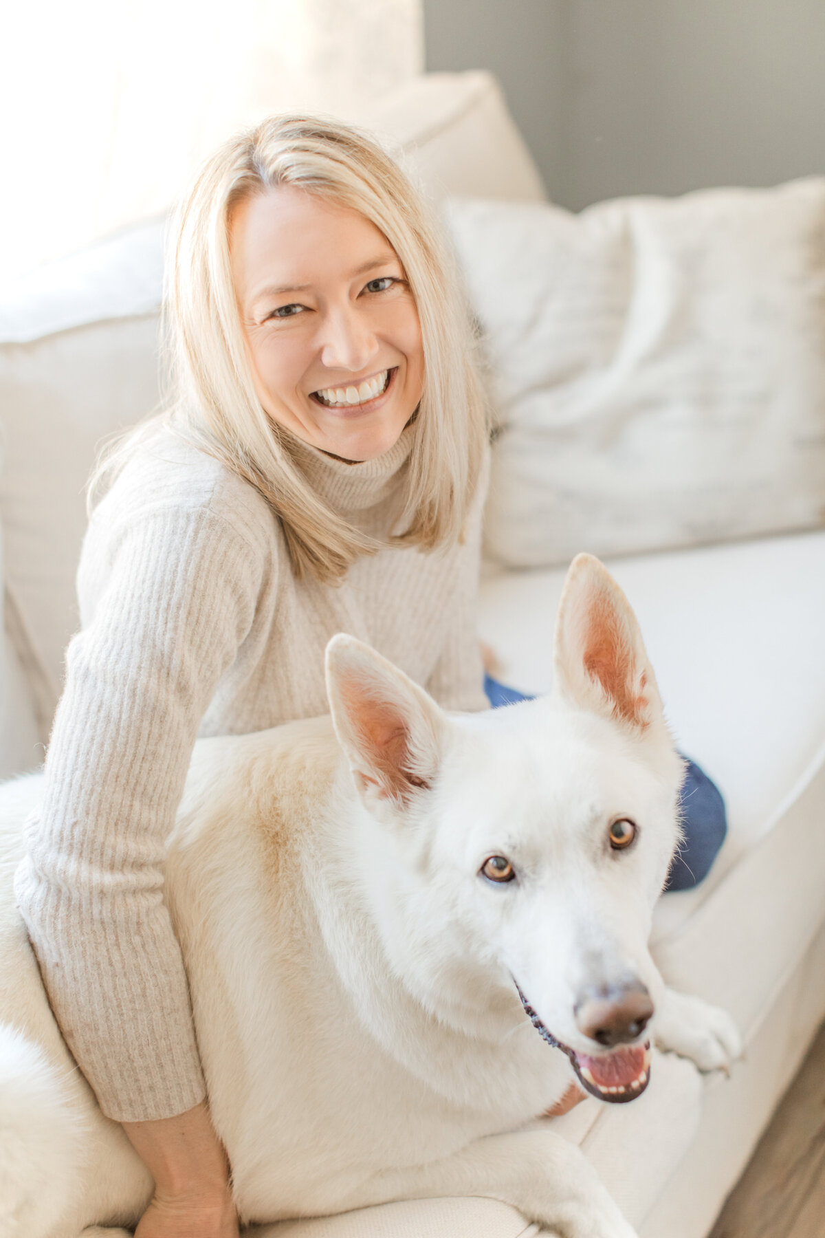 A joyful woman in a cream turtleneck sweater cuddles her white dog on a light sofa, showcasing warmth, comfort, and connection in a personal branding lifestyle session.