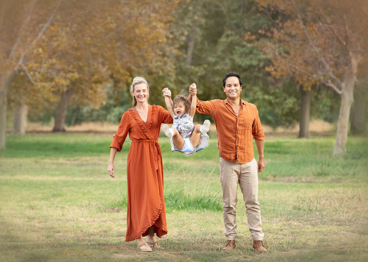 Mom and dad wearing burnt orange and swinging their daughter in the air in by Elsie Rose Photography
