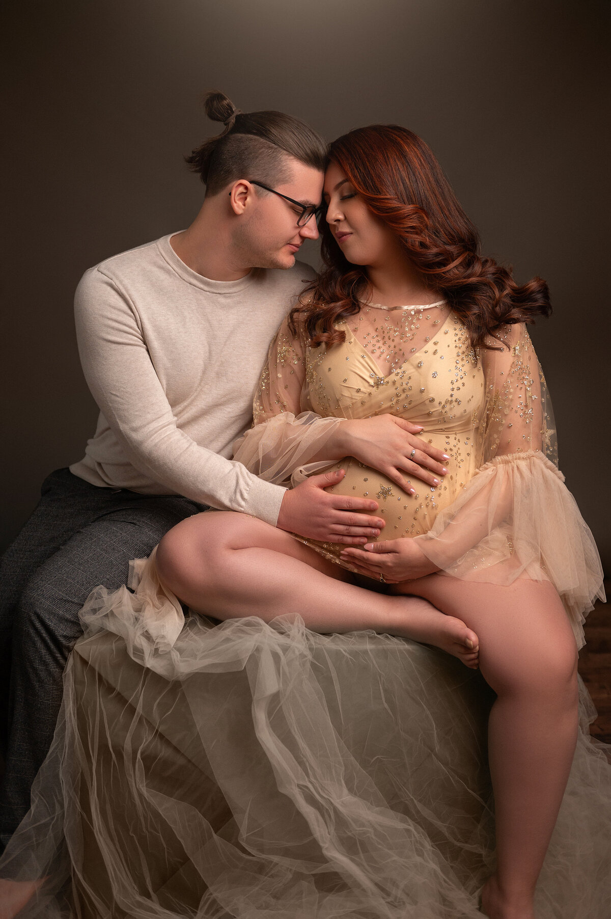 Pregnant woman in a sparkly gold tulle gown sitting close to her partner, foreheads touching, during an intimate and softly lit maternity session in Waukesha.