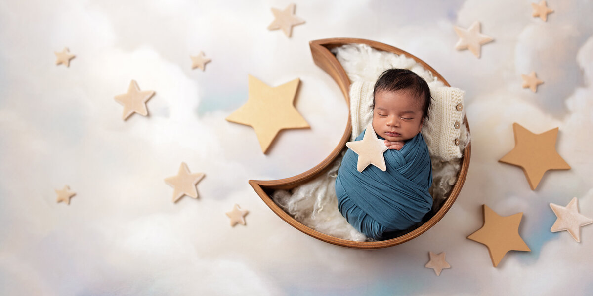 Newborn baby wrapped in blue and posed inside a wooden crescent moon prop surrounded by soft star accents on a cloudy sky background.