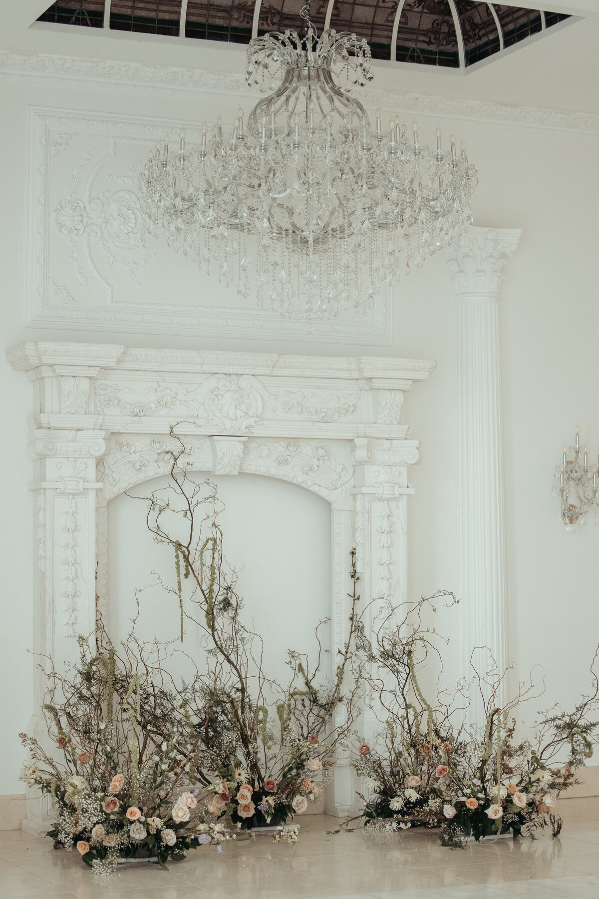 Ornate white fireplace with crystal chandelier and organic branch florals at an indoor Houston wedding.