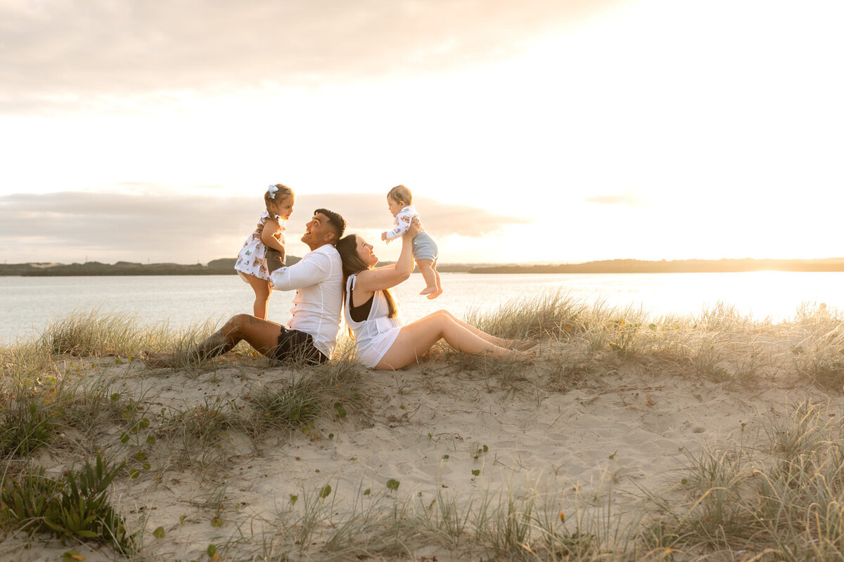 Young family enjoying a playful moment on sand dunes by the water at sunset, with parents holding their children in a joyful and light-filled setting.