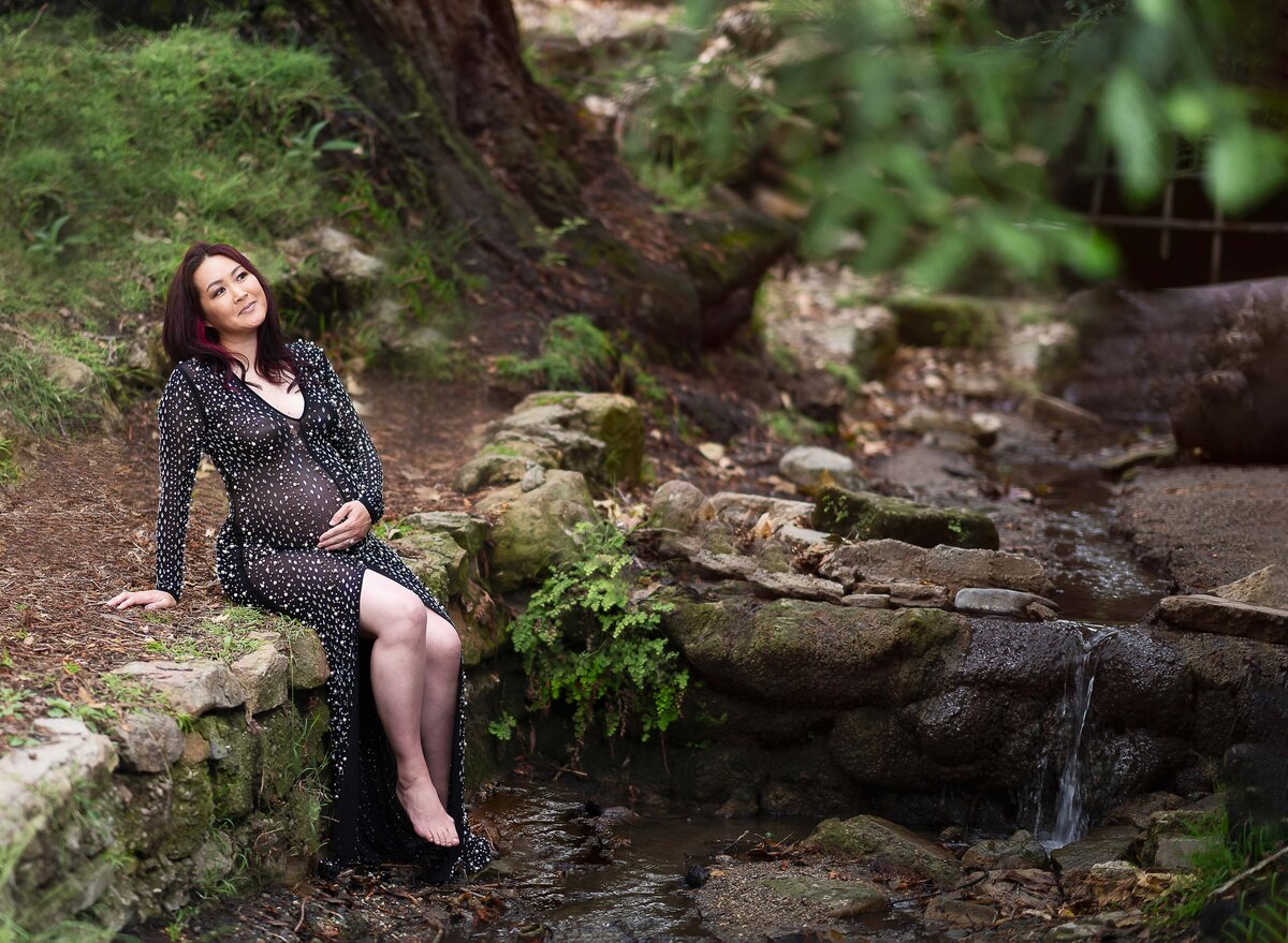 Mom in luxury maternity gown for outdoor maternity photoshoot in Philadelphia suburb forest. She is sitting on the edge of a creek with her toes touching the water, holding her baby bump and looking off in the distance.