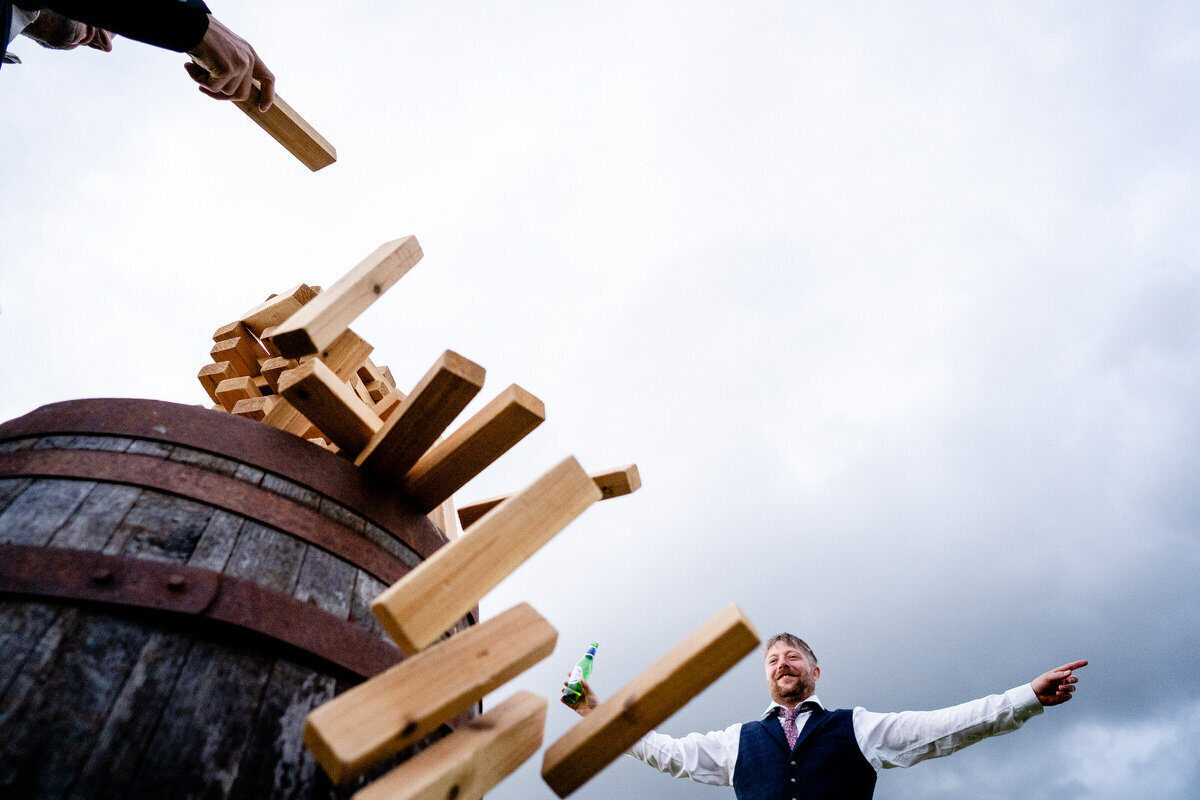 A wedding guest with his arms wide open celebrating with a beer in hand as he wins a game of jenga during an outdoor wedding in Gloucestershire 