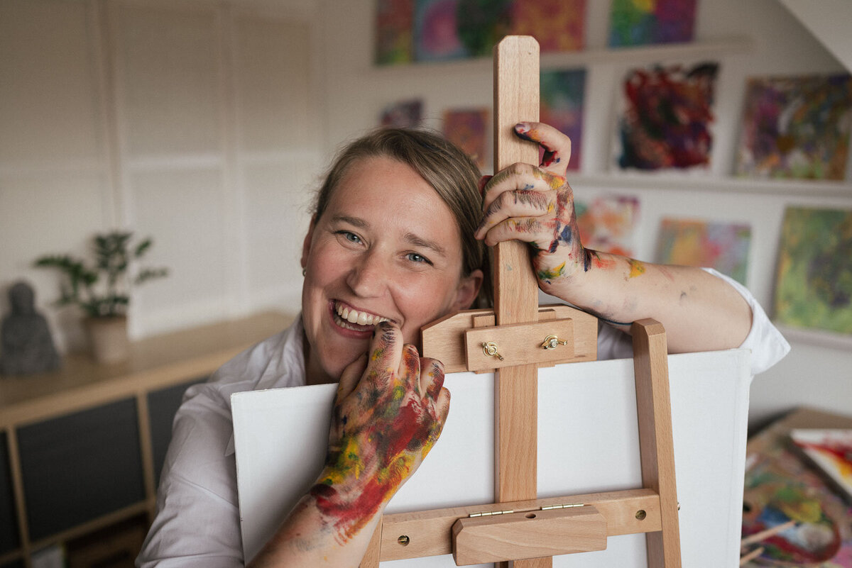 Künstlerin lacht hinter ihrer Staffellei in ihrem farbenfrohen Atelier