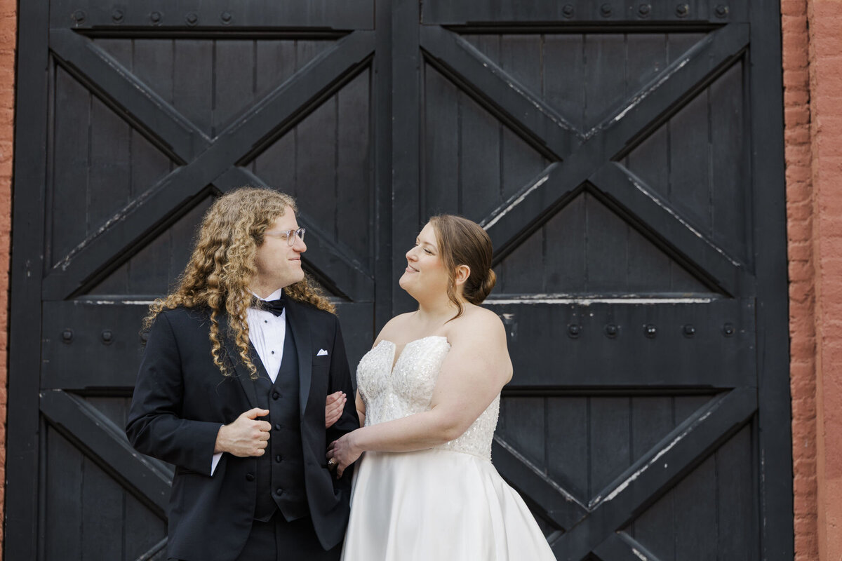 Two brides, one in a suit and one in a gown, standing in front of black barn doors at a Hudson Valley wedding