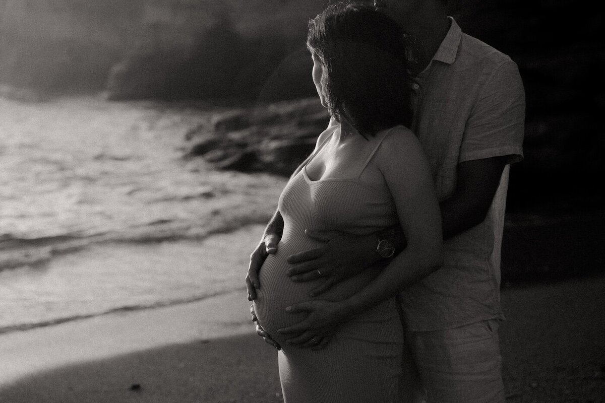 Le couple enlacé en bord de mer, les mains sur le ventre de la mère. Noir et blanc. Séance grossesse en Bretagne par Kamescop.