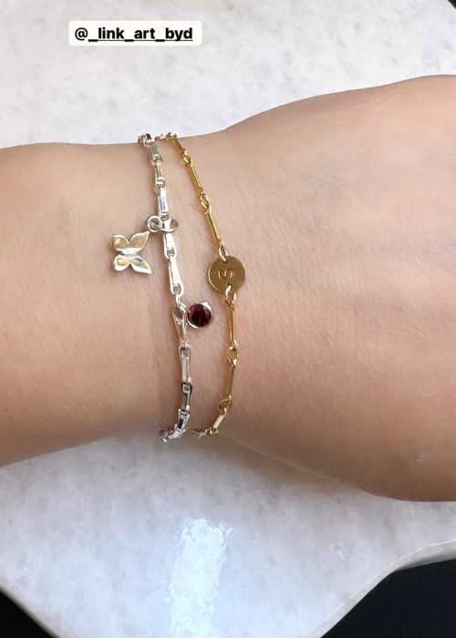 A hand displaying two charm bracelets—one silver and one gold—featuring butterfly, garnet, and initial charms from Link Art.