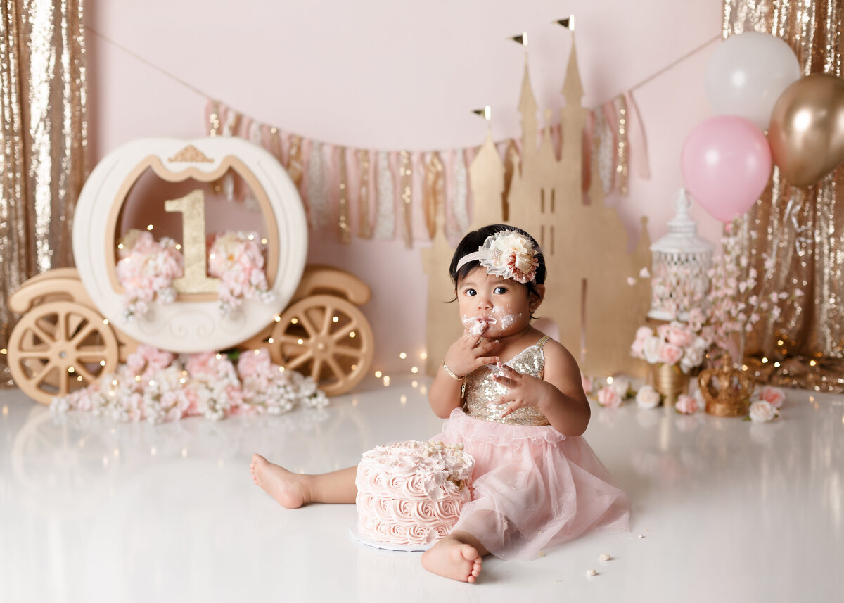 Cinderella themed cake smash at top West Palm Beach, FL newborn and cake smash photographer. Baby girl in a pink and gold dress is sitting in front of a pink rosette cake licking icing off of her fingers. In the background there is a white Cinderella cream and gold pumpkin carriage, gold castle cutout, flowers, and balloons.