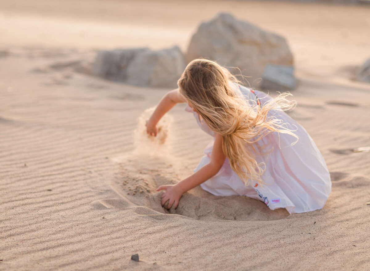 Sweet moment of a young girl with long hair playing and digging in the sand at the beach, captured by Leslie Q Photography
